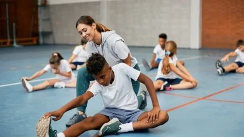 A smiling female physical education teacher or fitness trainer is kneeling and assisting a young Black boy with a seated toe-touch stretch on a blue gymnasium floor. Several other diverse children are visible in the background performing the same stretch.