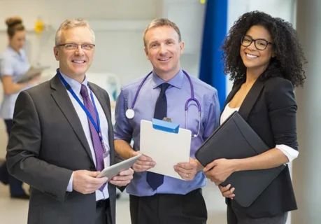 A group of healthcare professionals, including a doctor with a stethoscope and administrators in business attire, smiling in a hospital setting, illustrating roles in nursing and healthcare administration.