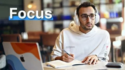 A thumbnail-style image for a video or article, featuring a young man with glasses and a white hoodie sitting at a table in a bright, blurry cafe or library setting. He is wearing white over-ear headphones, looking intensely at something off-camera, and actively writing in a notebook with a pen. A laptop with an Apple logo is visible to his left. The word "Focus" is prominently displayed in large white text with a blue background shadow in the upper left corner.