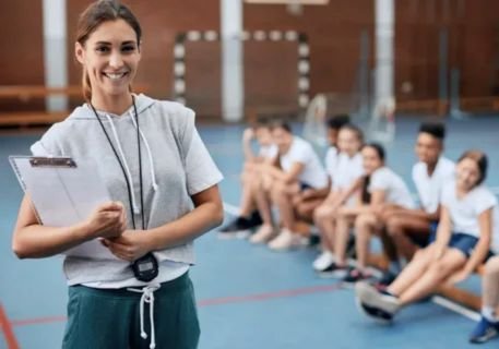 A smiling female physical education teacher or coach, wearing a gray hooded shirt and a stopwatch around her neck, stands in a gymnasium holding a clipboard. In the blurred background, a line of diverse students, wearing white shirts and shorts, is seated on a bench or bleacher.