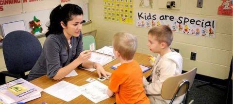 The image shows a female Special Education teacher working individually with two young male students at a table in a classroom setting.
The teacher is holding a card, likely a visual aid or flashcard, while the students look on and interact with materials spread on the table. The interaction appears to be a focused small-group or one-on-one lesson.